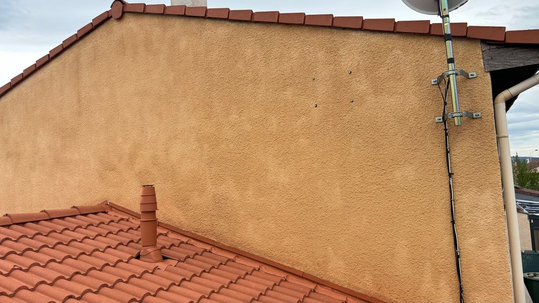 Mur en stuc beige, toit de tuiles rouges et antenne parabolique sur fond de ciel nuageux.