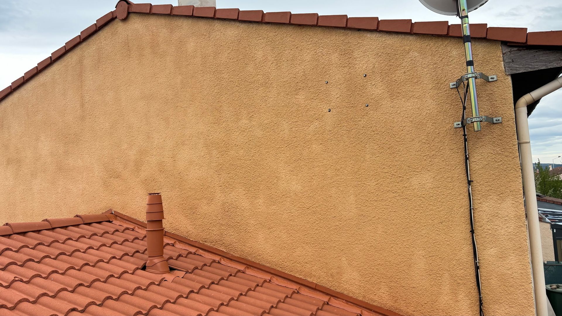 Mur en stuc beige, toit de tuiles rouges et antenne parabolique sur fond de ciel nuageux.