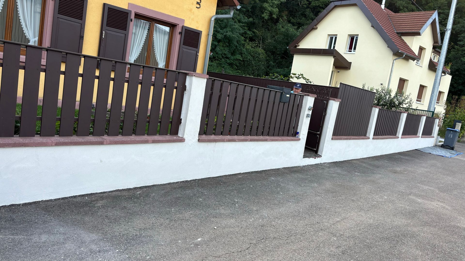 Clôture en bois brun sur un mur blanc, avec un petit portail et deux maisons jaunes en arrière-plan.