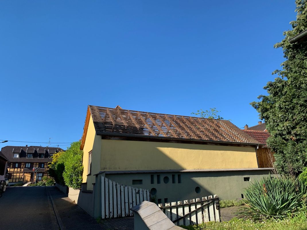 Bâtiment jaune au toit délabré se détachant sur un ciel bleu. Clôture blanche devant.