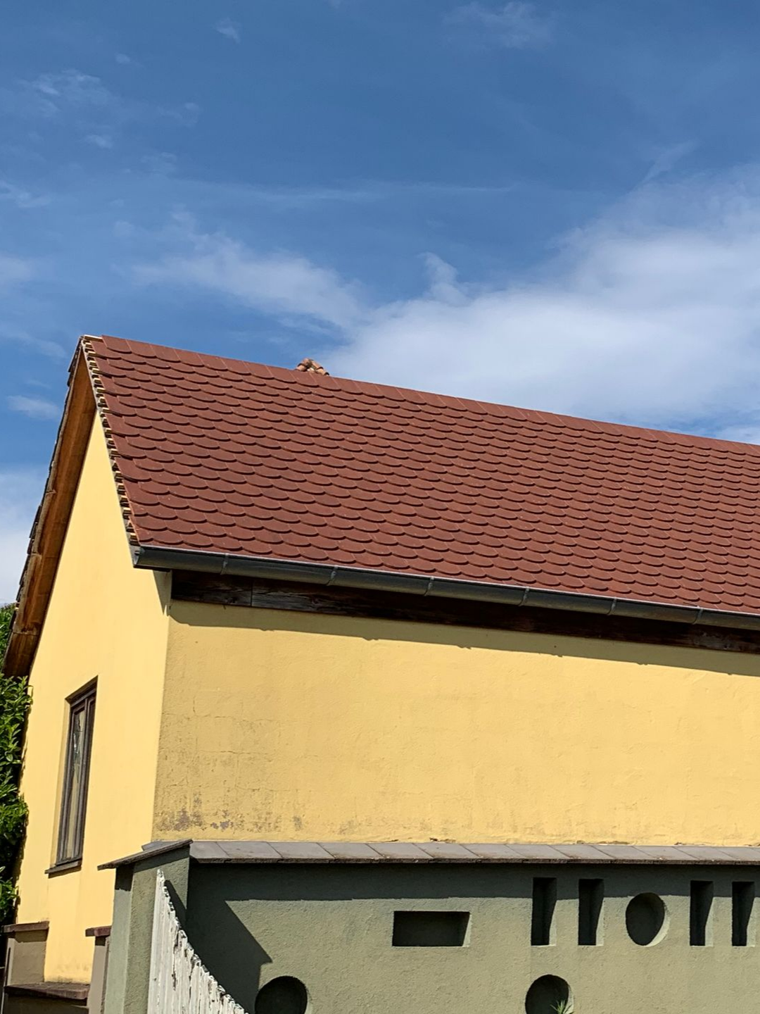 Bâtiment jaune au toit de tuiles rouges se détachant sur un ciel bleu. Un muret en béton est visible au premier plan.