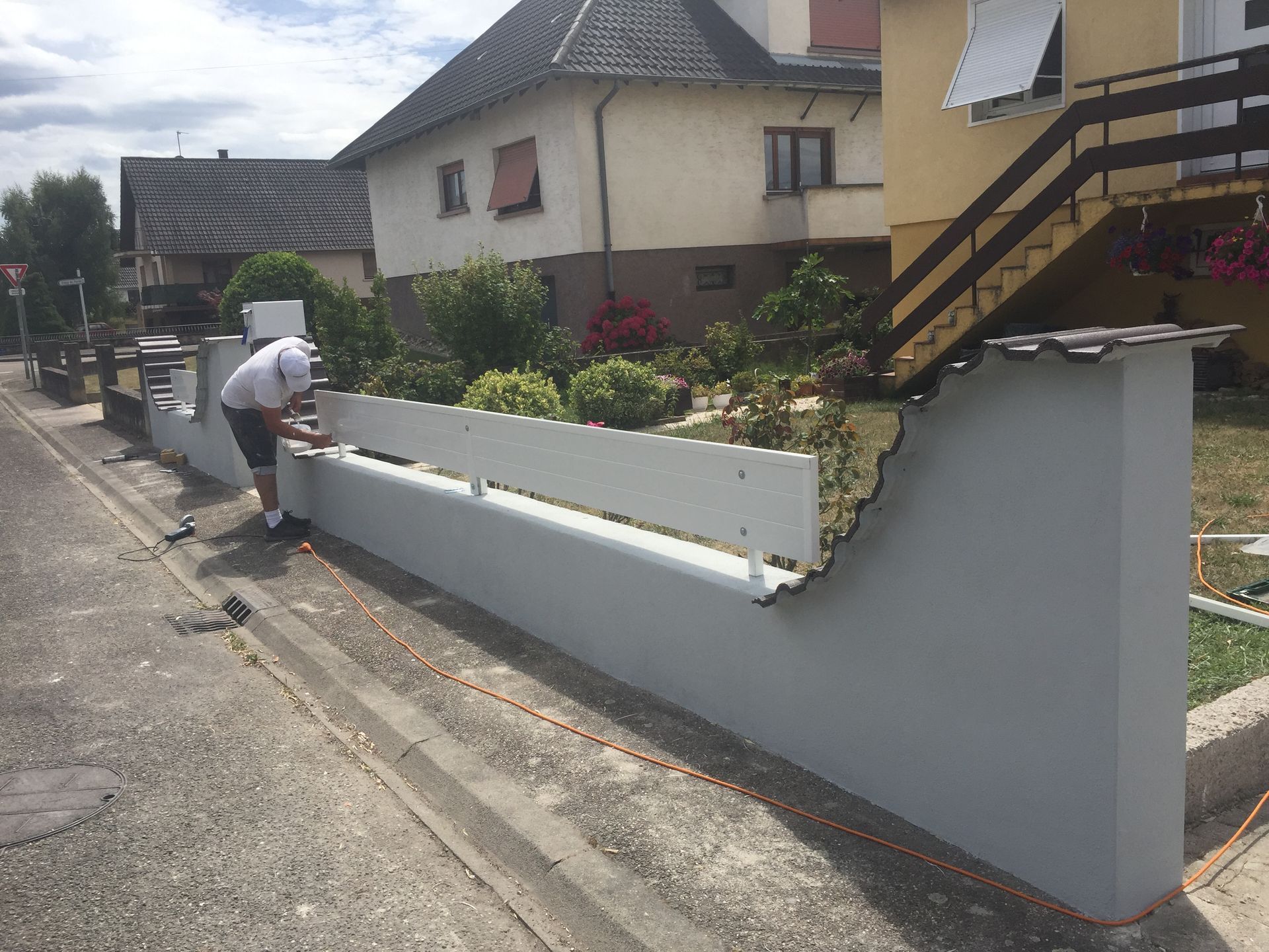 Une personne installe une clôture blanche sur un mur gris au bord d'une route ; des maisons sont visibles en arrière-plan.