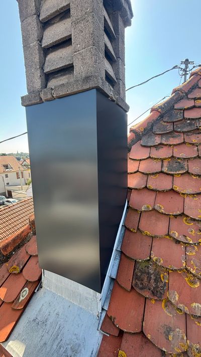 Couvercle de cheminée en métal noir sur un toit de tuiles rouges, à côté d'une cheminée en pierre, au soleil.