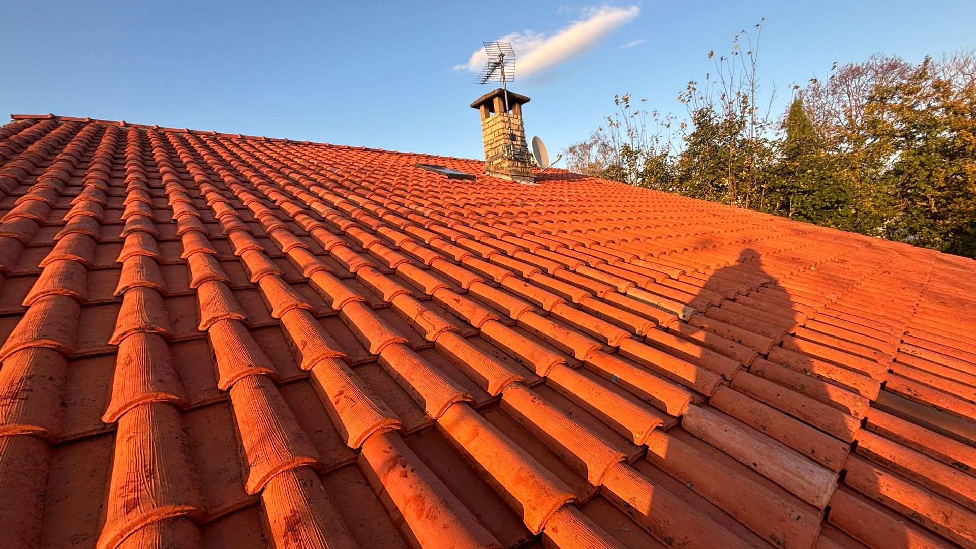 Toit de tuiles rouges avec cheminée et girouette sur fond de ciel bleu.