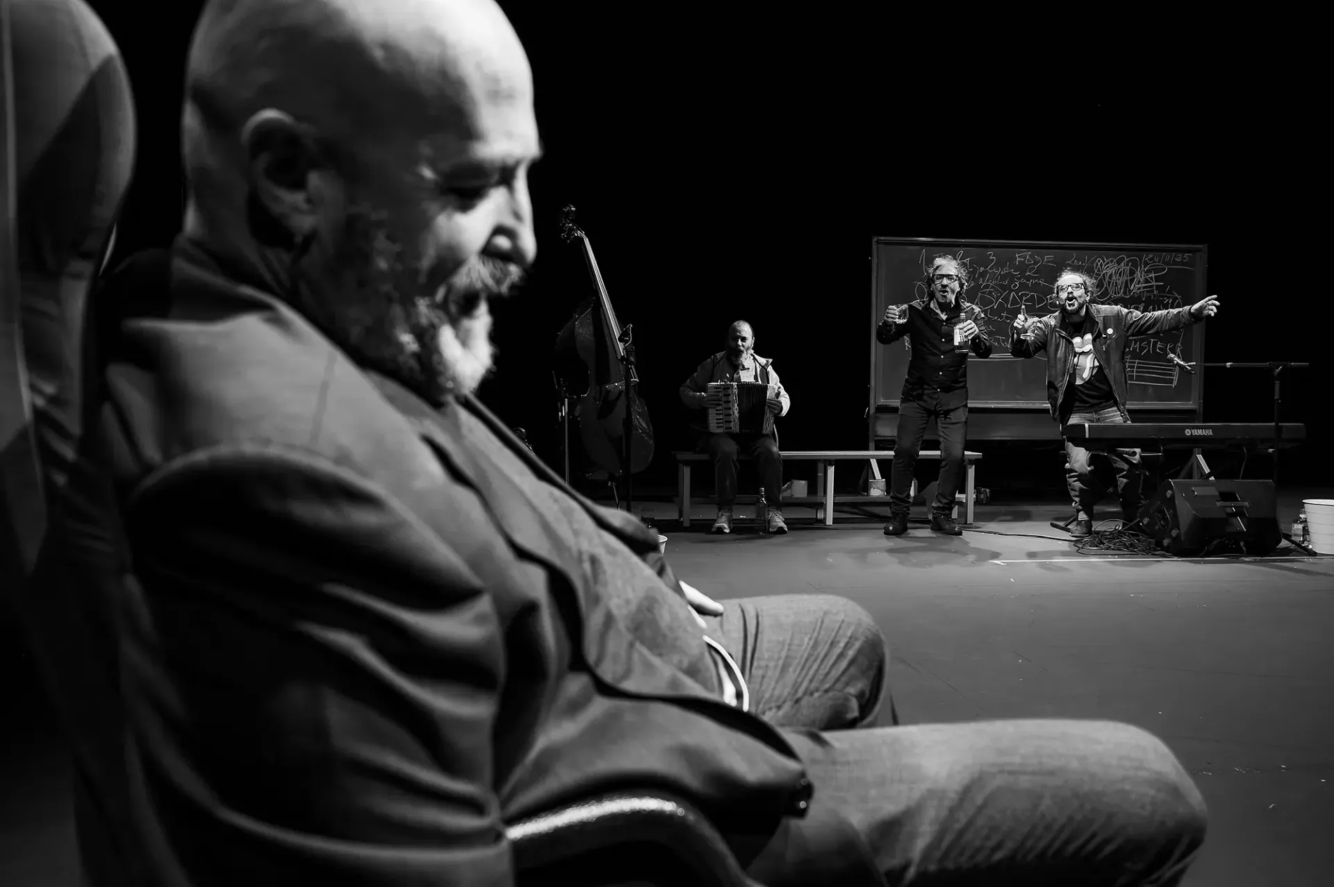 Fotografía en blanco y negro: Un hombre con una chaqueta observa a una banda tocar en el escenario.