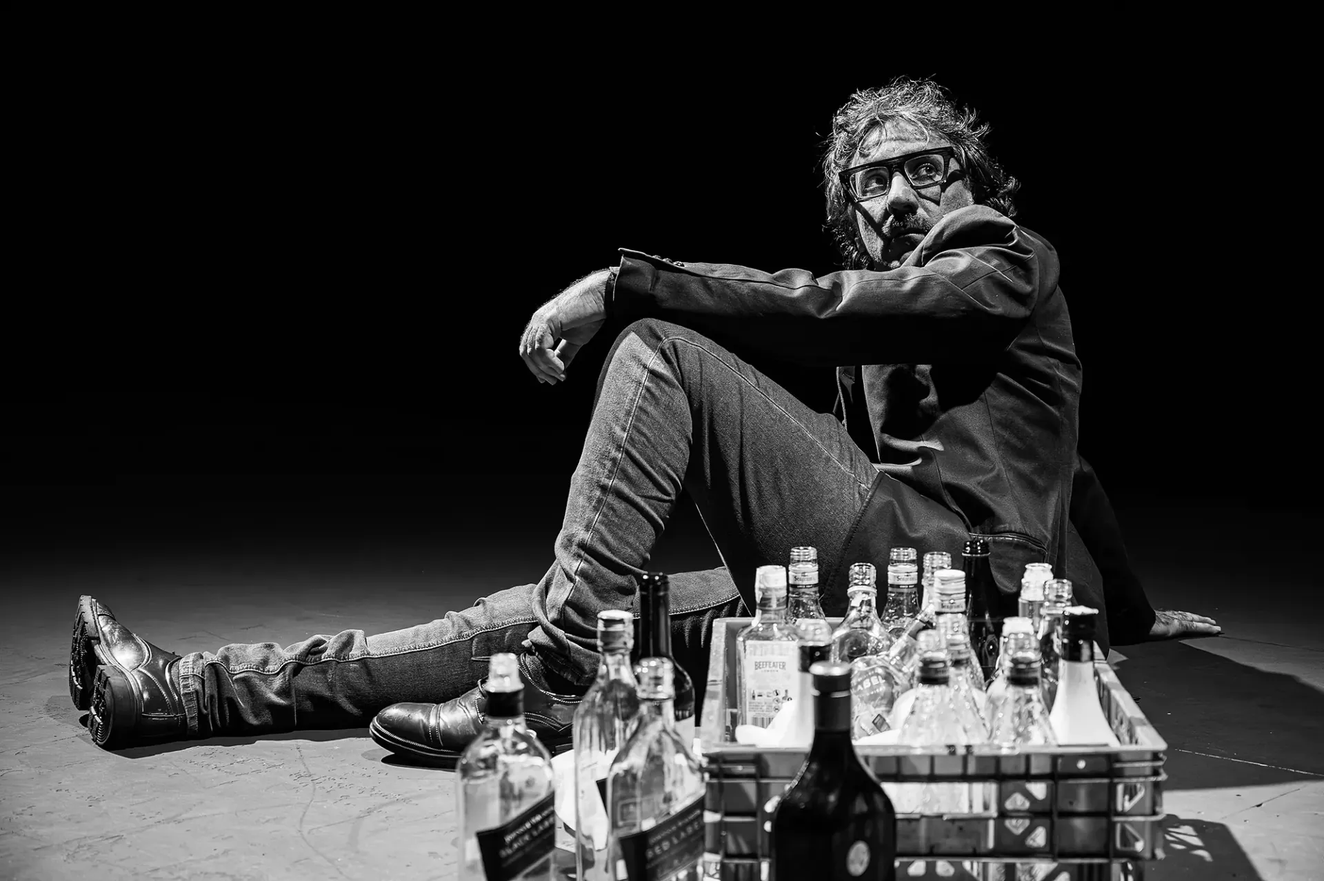 Un hombre con chaqueta está sentado en el suelo junto a una caja de botellas de licor. Foto en blanco y negro.