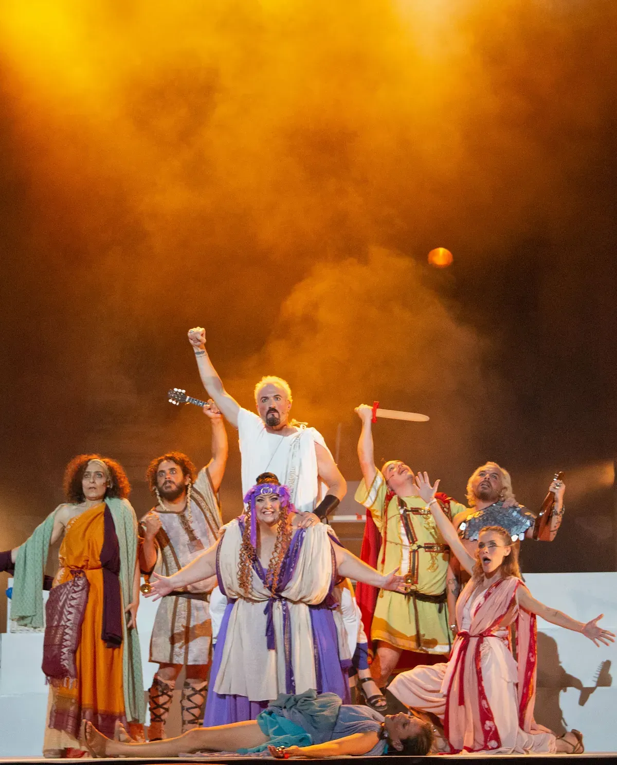 Reparto teatral con trajes de la antigua Roma, actuando en escena. Fondo amarillo, espadas y brazos en alto.