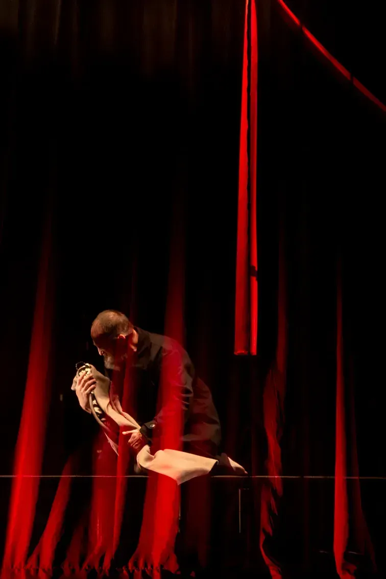 Una persona vestida de oscuro se arrodilla en un escenario, sosteniendo un objeto blanco, iluminado por luces rojas.