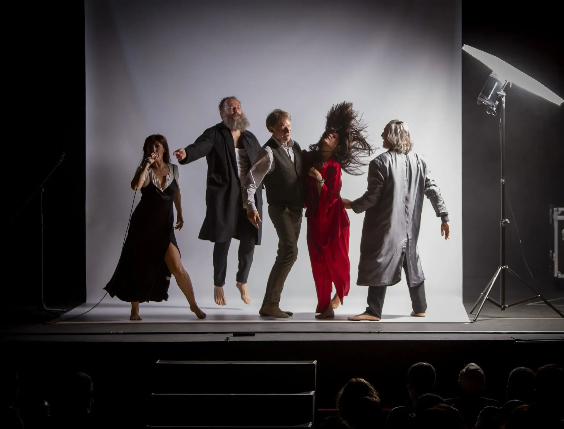 Representación teatral: cinco actores en escena. Uno canta, otros visten abrigos o vestidos, con diversas expresiones.