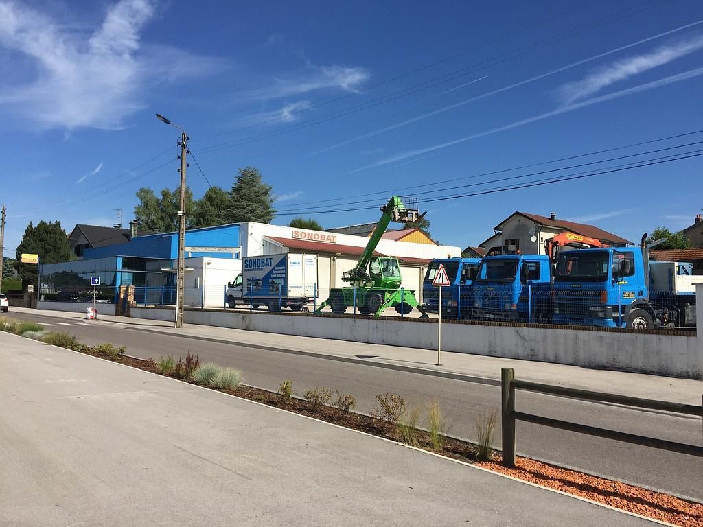 Extérieur d'une entreprise avec des camions bleus, un monte-charges vert et un bâtiment blanc sous un ciel bleu.