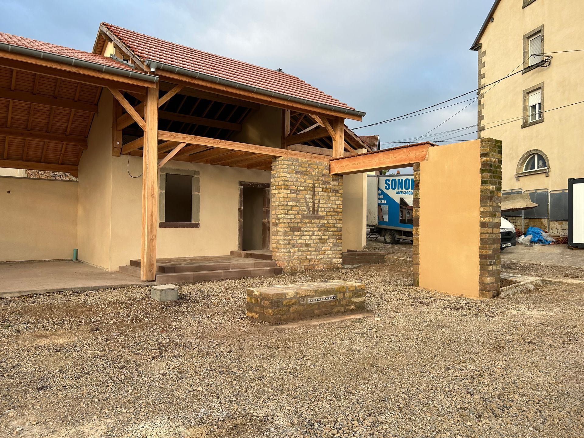 Extérieur du bâtiment en construction avec poutres en bois apparentes, murs en pierre et couleur beige, et sol en gravier.