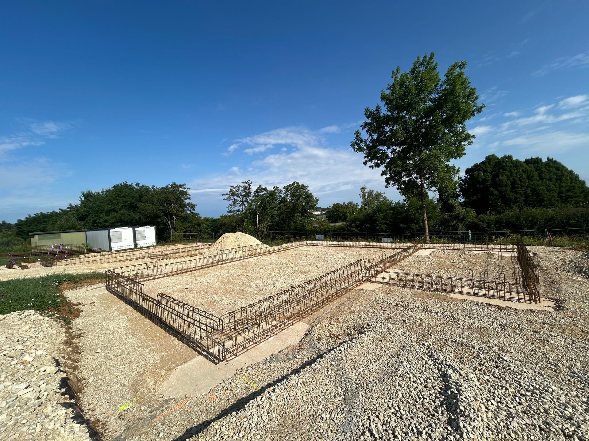 Chantier de construction avec fondations en barres d'armature apparentes sur gravier, arbre en arrière-plan, ciel bleu.