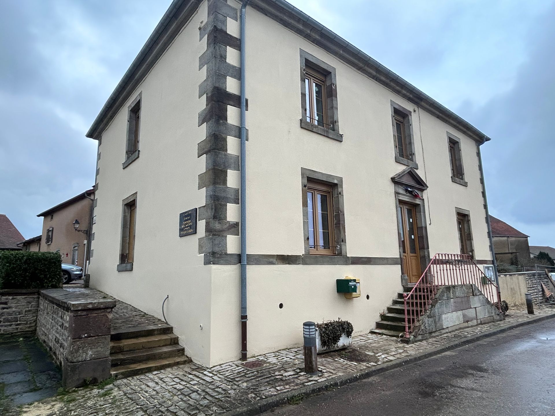 Bâtiment de deux étages avec des ornements en pierre, des fenêtres et une porte marron, et des marches menant à l'entrée, par temps nuageux.
