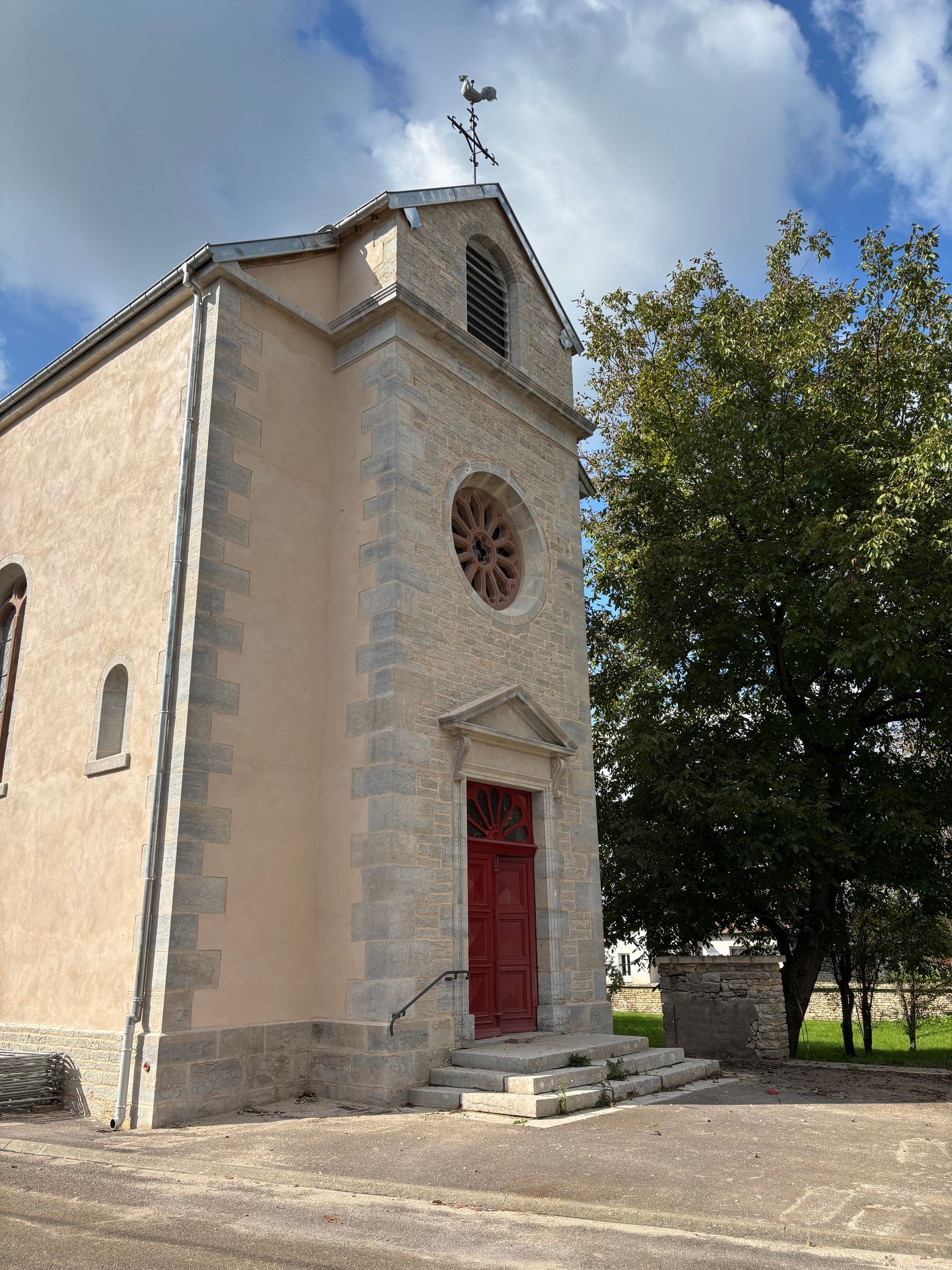 Maison en pierre avec volets verts, arbre et pelouse avec tables blanches et chaises rouges.