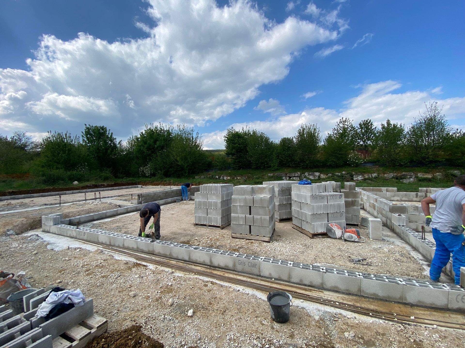 Chantier de construction avec des ouvriers posant des blocs de béton gris ; ciel bleu nuageux.