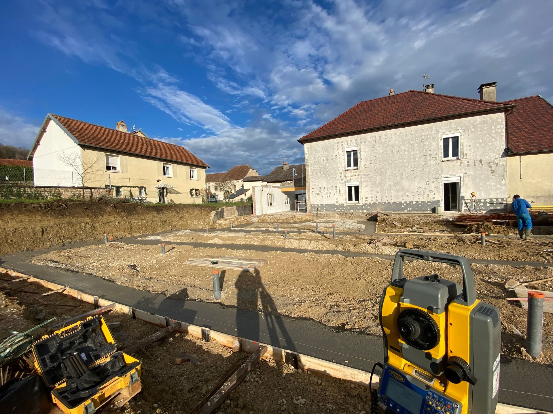 Chantier de construction avec fondations, outils et deux bâtiments sous un ciel bleu.