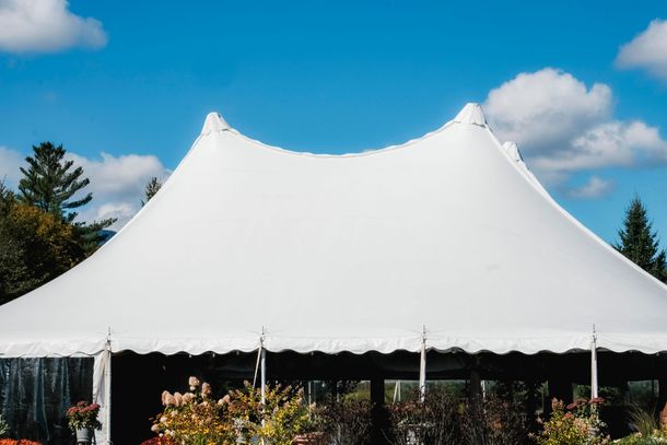 Grande tente blanche événementielle à toit pointu se détachant sur un ciel bleu parsemé de nuages, entourée de verdure et de fleurs.
