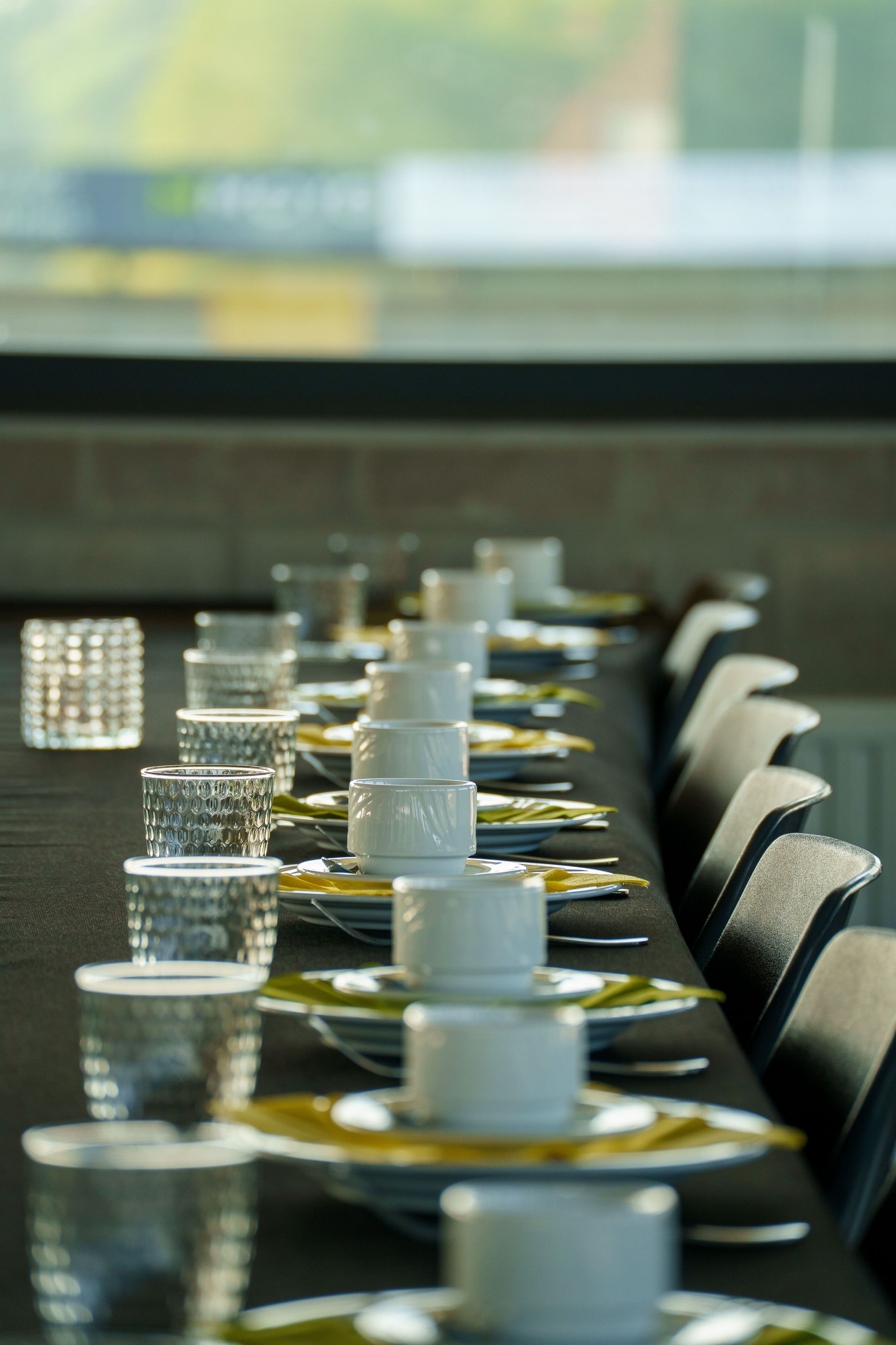 Table à manger soigneusement dressée avec tasses blanches, soucoupes et verres transparents près de la lumière naturelle.