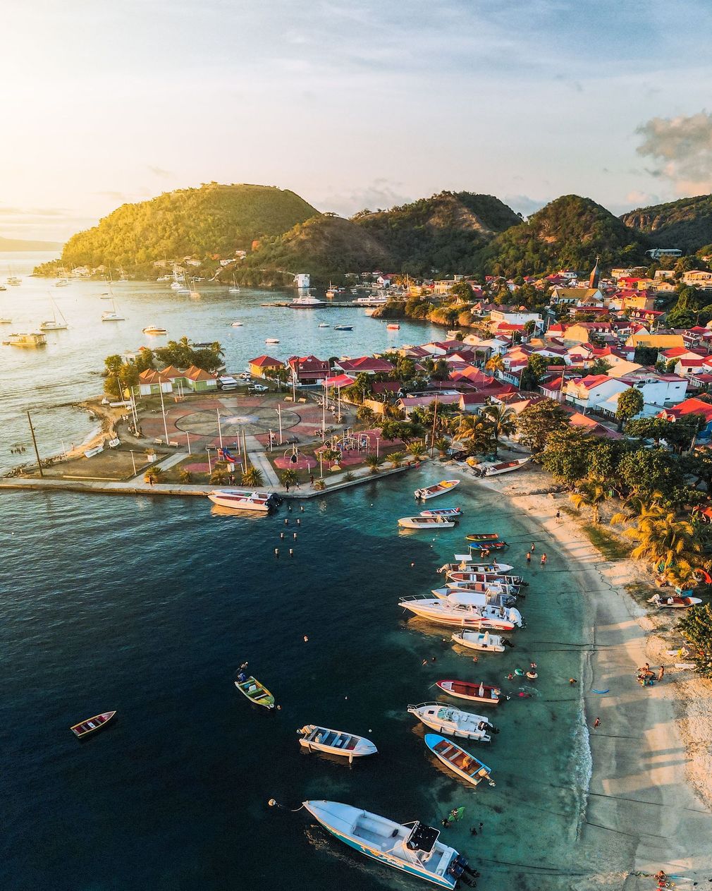 Vue aérienne d'une ville balnéaire tropicale aux toits colorés, des bateaux dans une baie turquoise et un rivage illuminé par le coucher du soleil.