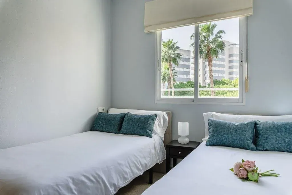 Dormitorio con dos camas, sábanas blancas, almohadas color verde azulado, ventana con palmeras, paredes azul claro.