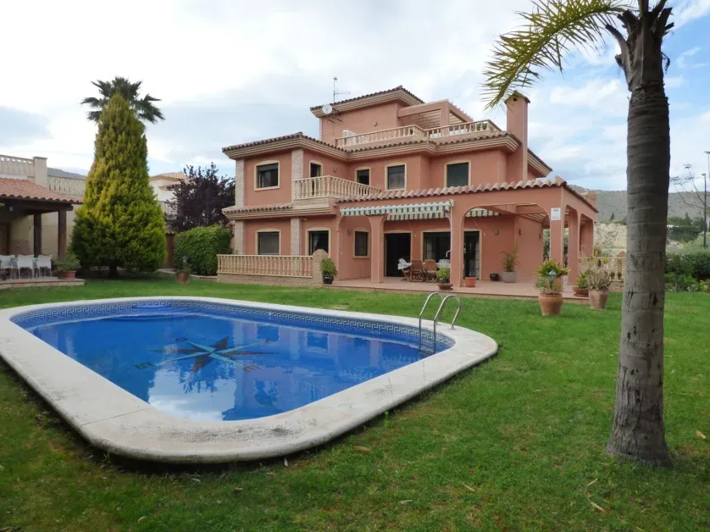 Casa grande color melocotón con piscina en el patio delantero. Césped verde, palmeras y cielo despejado.