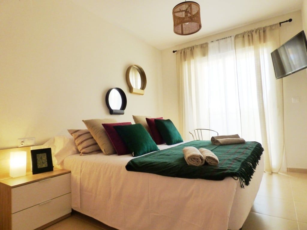 Dormitorio con ropa de cama blanca, manta verde, cojines, espejos y un televisor montado en la pared.