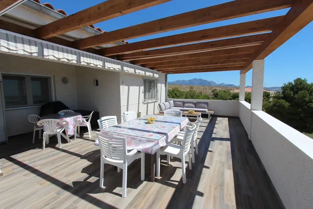 Patio al aire libre con mesa de comedor y asientos bajo una pérgola de madera, con vistas a un paisaje.