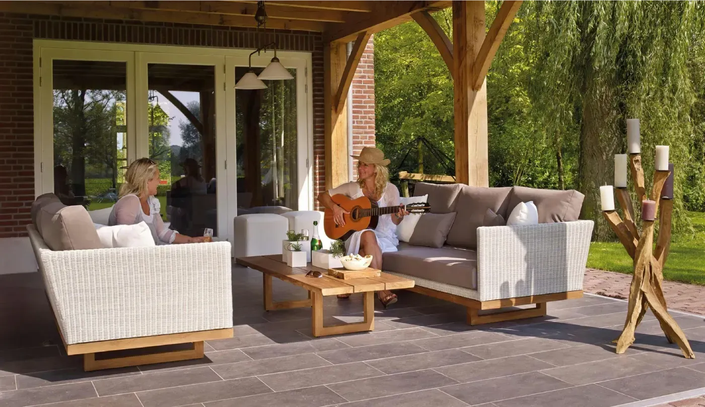 Dos personas en sofás de mimbre al aire libre, una tocando la guitarra. Patio con mesa, velas y árboles.