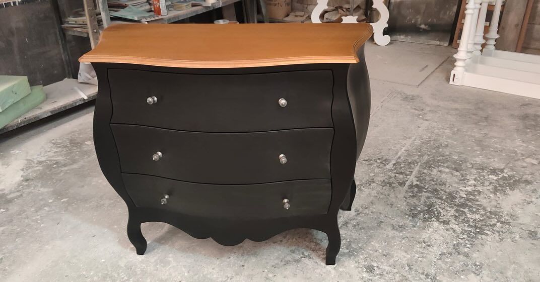 Cómoda antigua de color negro y marrón con tres cajones, tiradores plateados y frente curvo, colocada sobre el suelo de un taller.