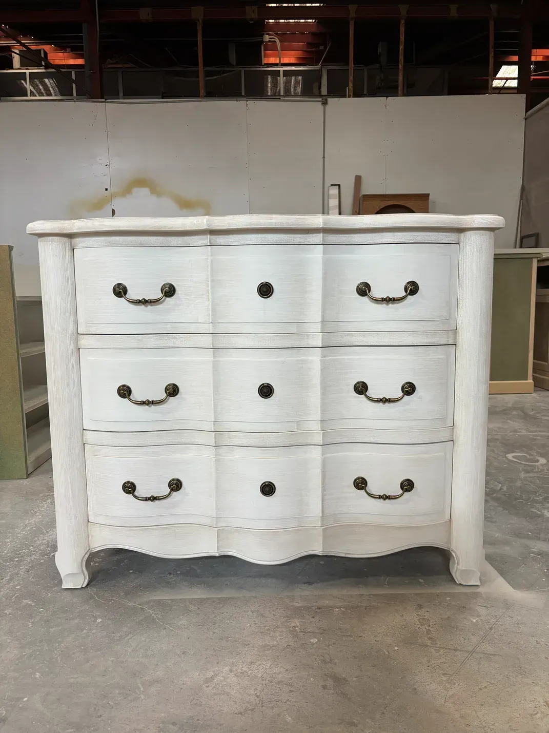 Cómoda de tres cajones en color blanco desgastado con tiradores de color negro, en un entorno de almacén.