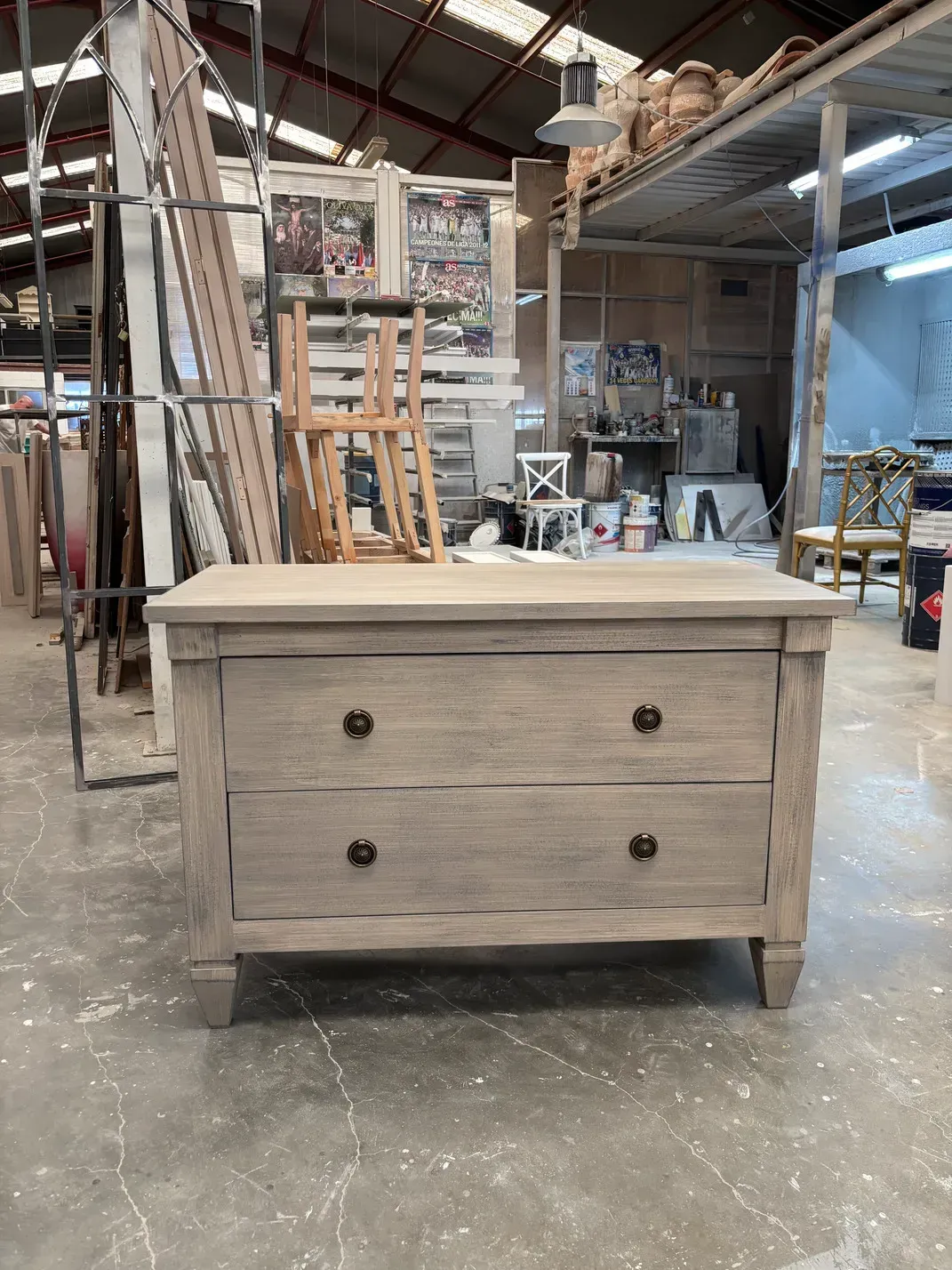 Cómoda de madera de dos cajones con tiradores oscuros en un taller, otros muebles al fondo.
