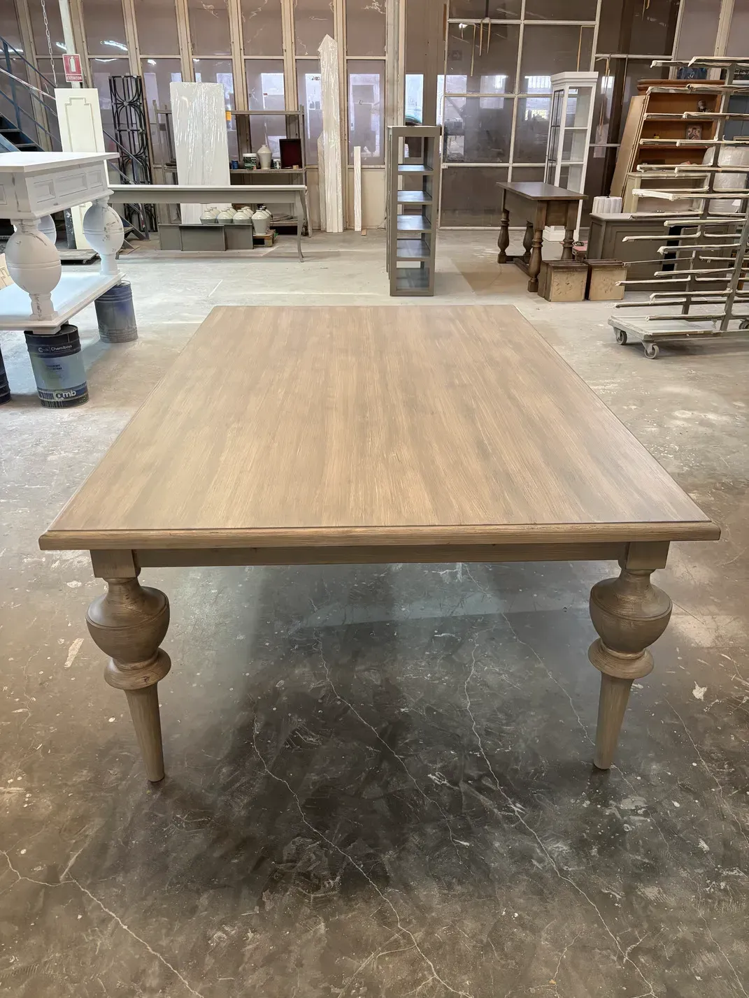 Gran mesa de comedor de madera clara con patas torneadas, en entorno de taller.