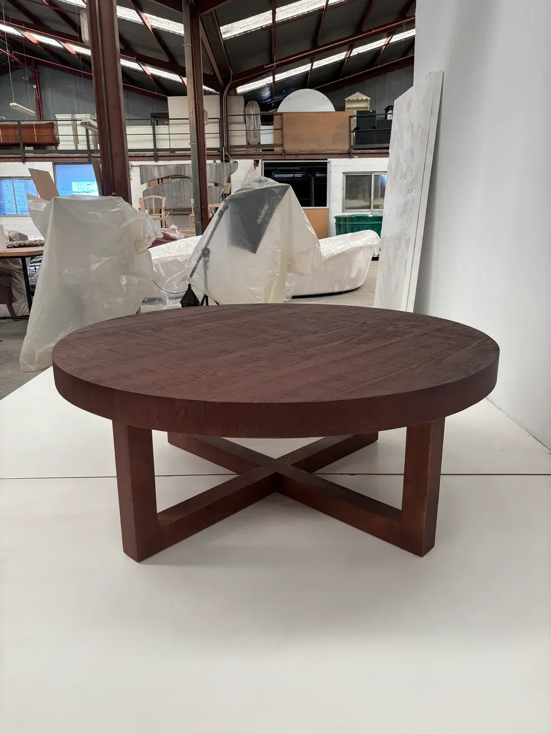 Mesa de centro redonda marrón con patas entrecruzadas en un entorno de almacén.