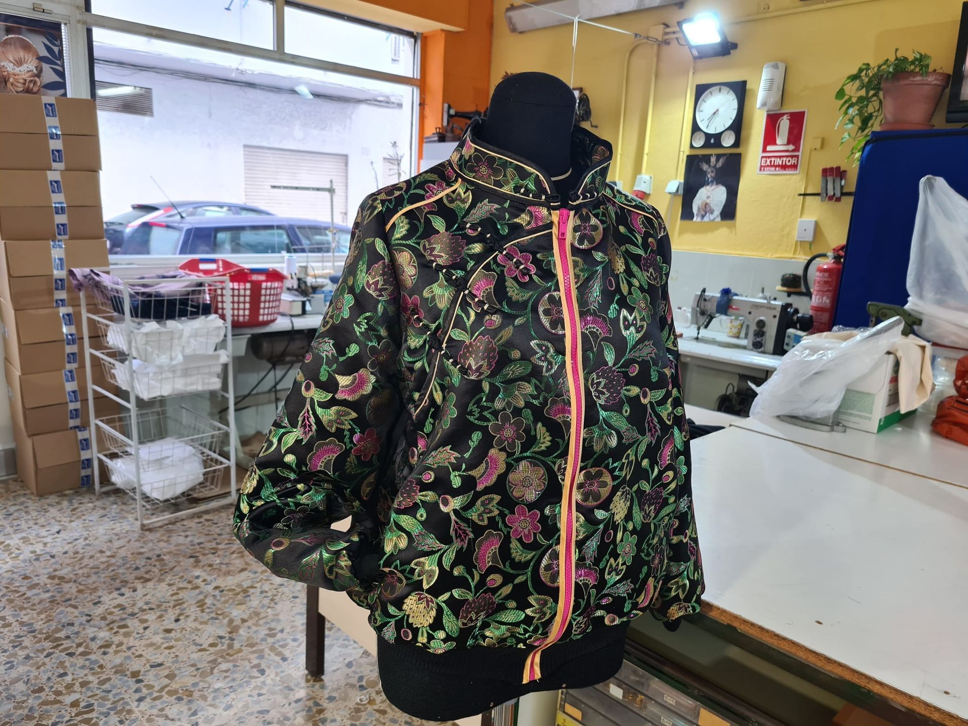 Chaqueta floral negra sobre maniquí en un entorno de taller con máquina de coser y ventana visibles.