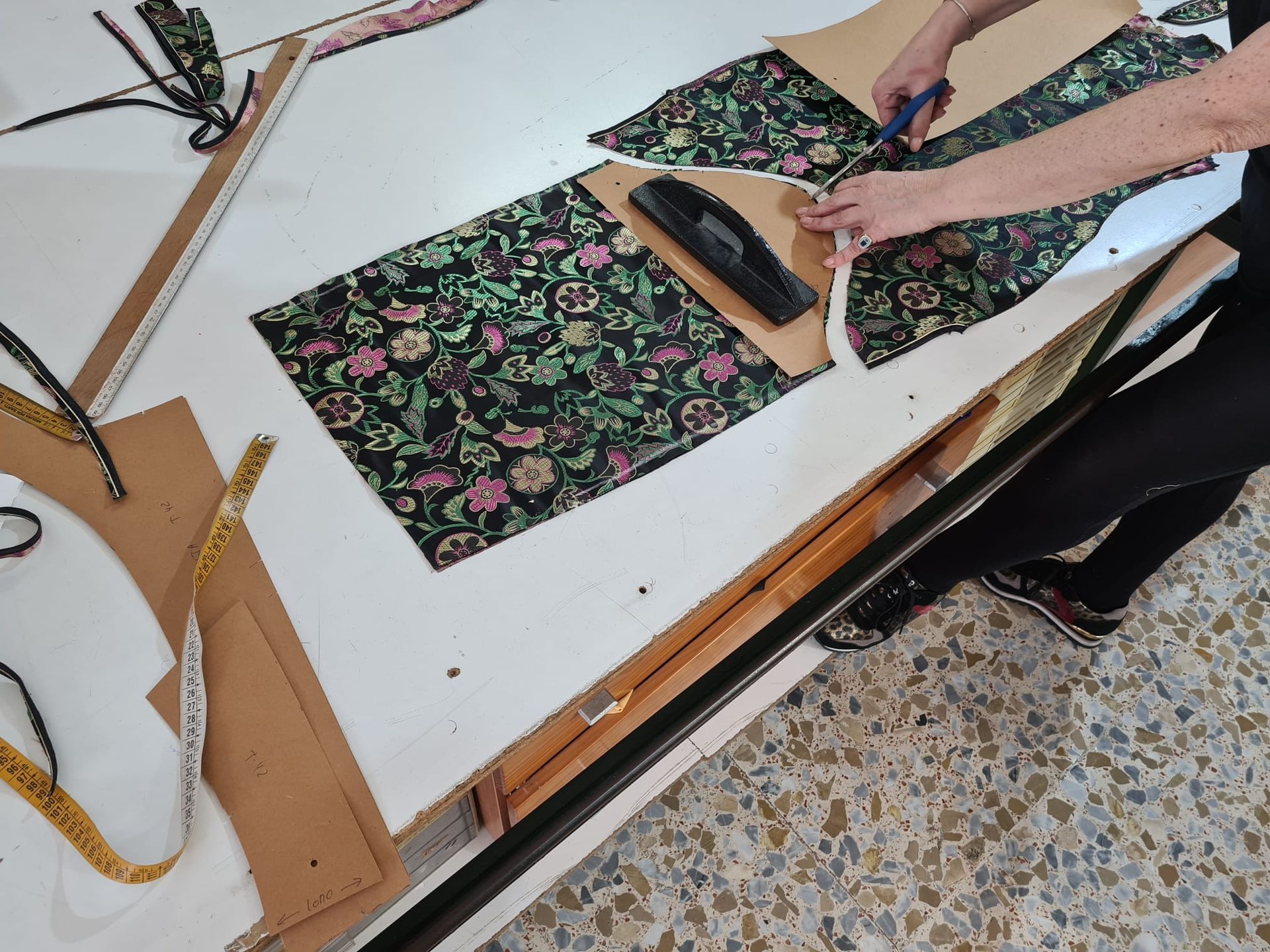 Persona cortando tela con tijeras usando un patrón sobre una mesa blanca. Tela floral, herramientas de costura.