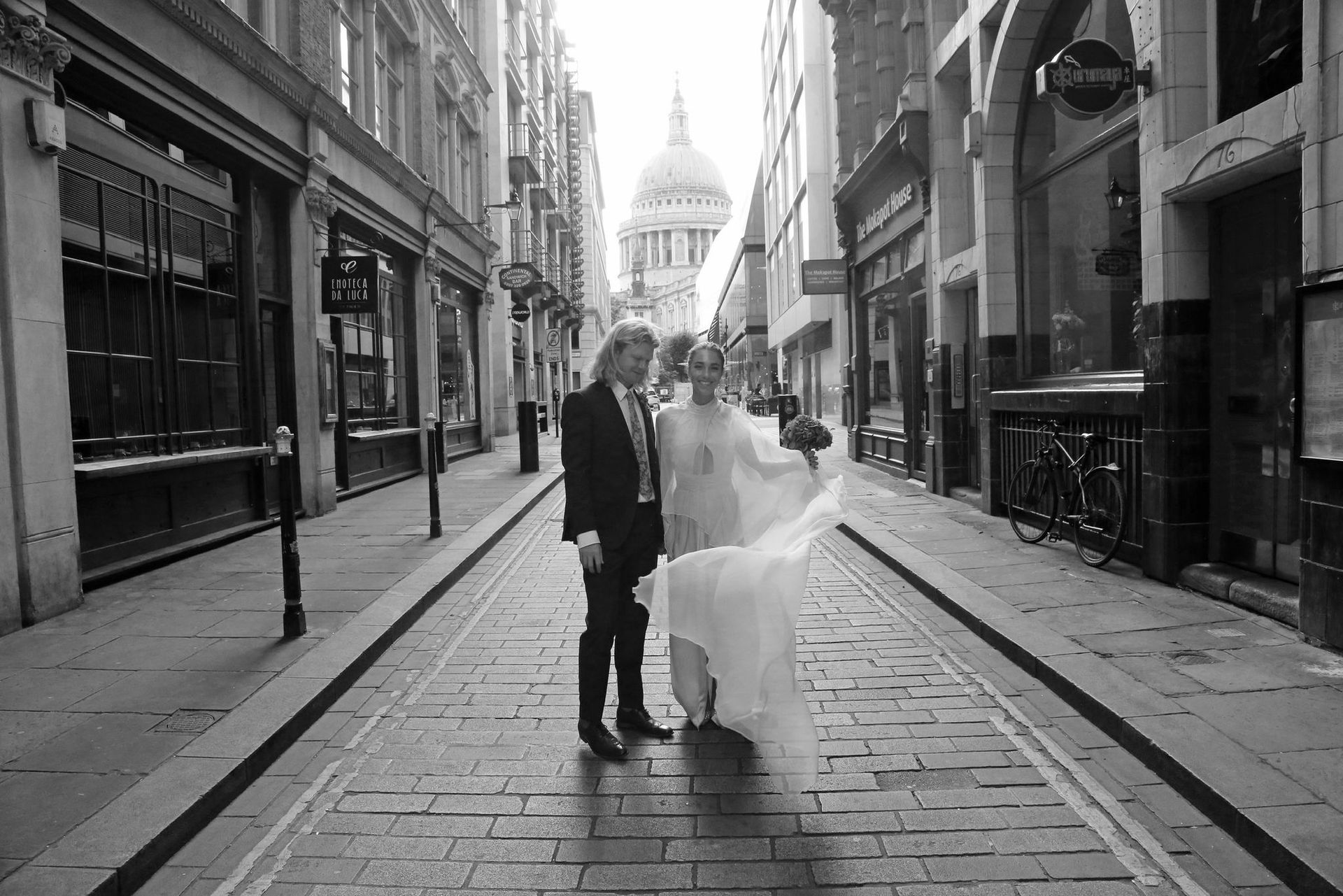 Novios posan en una calle adoquinada, con la Catedral de San Pablo al fondo. Blanco y negro.