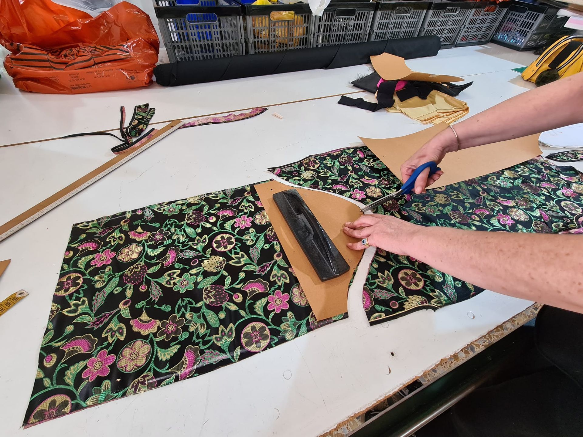 Manos cortando tela con tijeras y piezas de patrón sobre una mesa blanca.