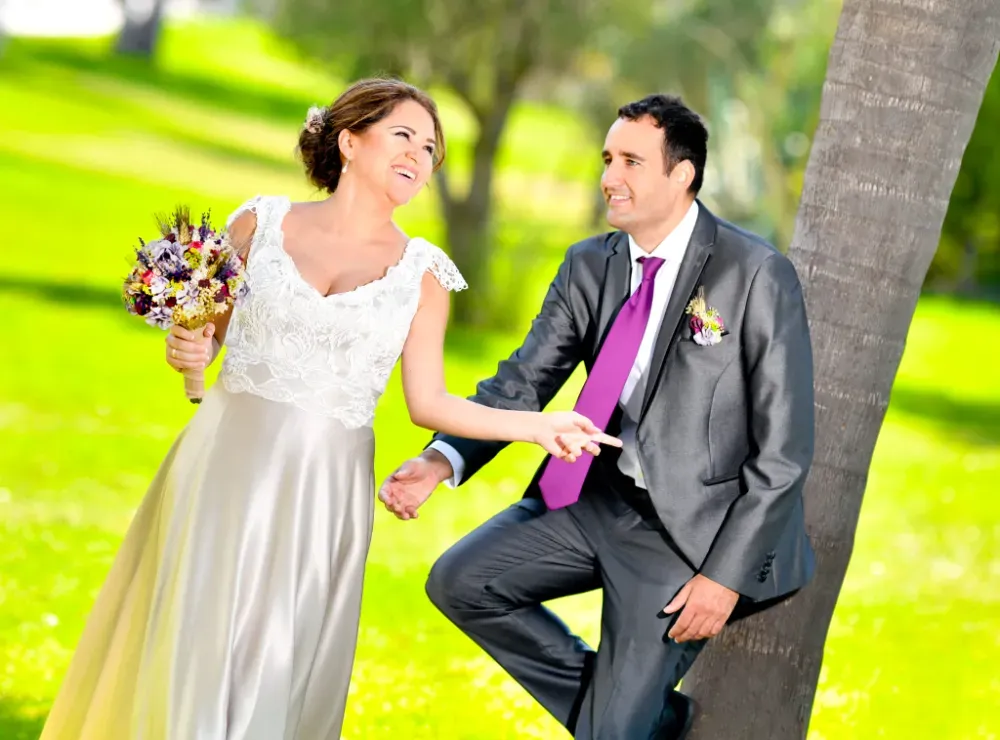 Novia y novio riendo, tomados de la mano cerca de un árbol, en un parque soleado; ella lleva un vestido blanco, él lleva un traje.