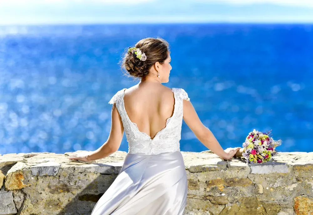 Novia con vestido de novia, mirando hacia el océano. Sostiene un ramo de flores y se apoya en un muro de piedra, con el agua azul como fondo.