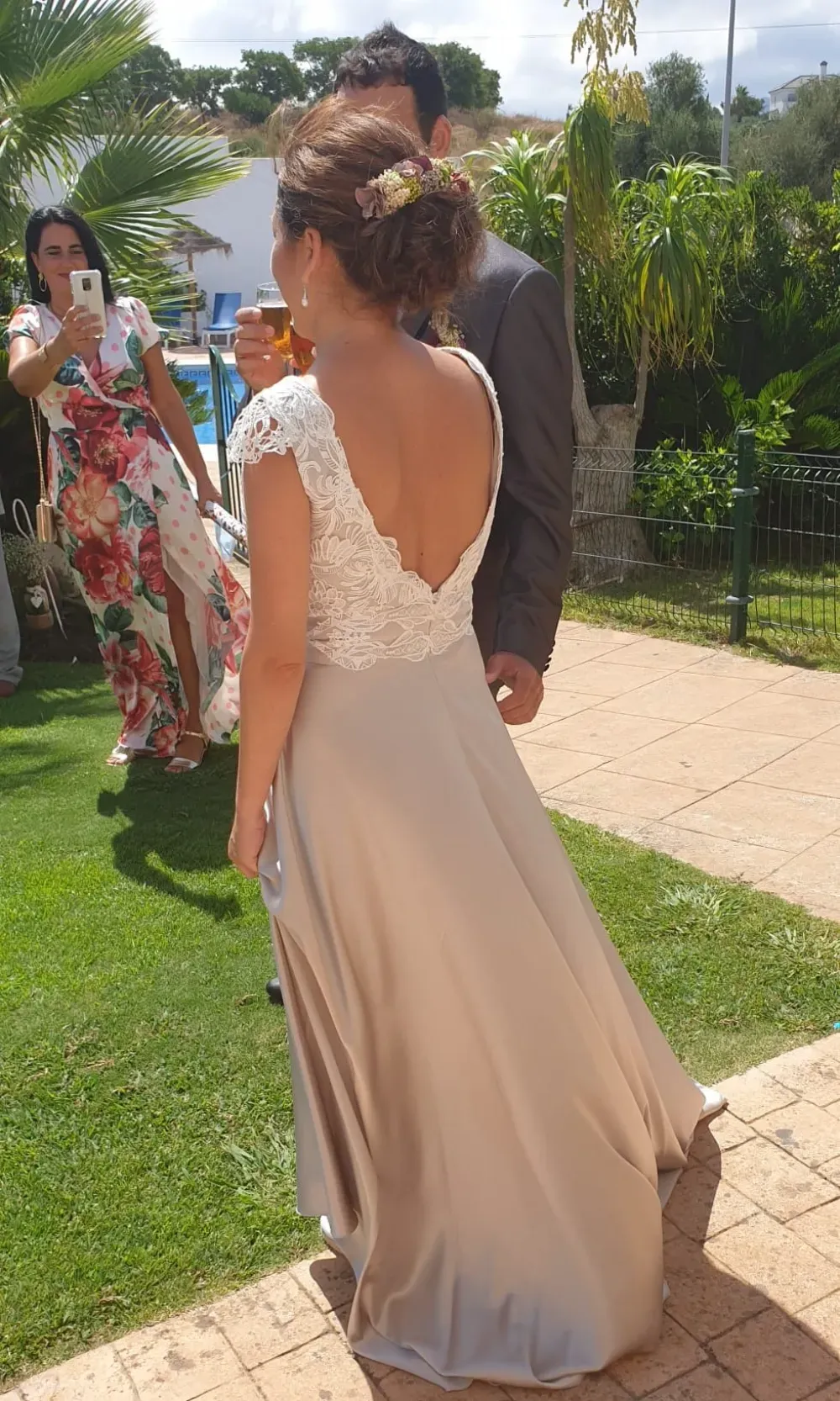 Mujer con vestido beige y corpiño de encaje, con la espalda al descubierto. Invitados junto a la piscina y un hombre con traje.