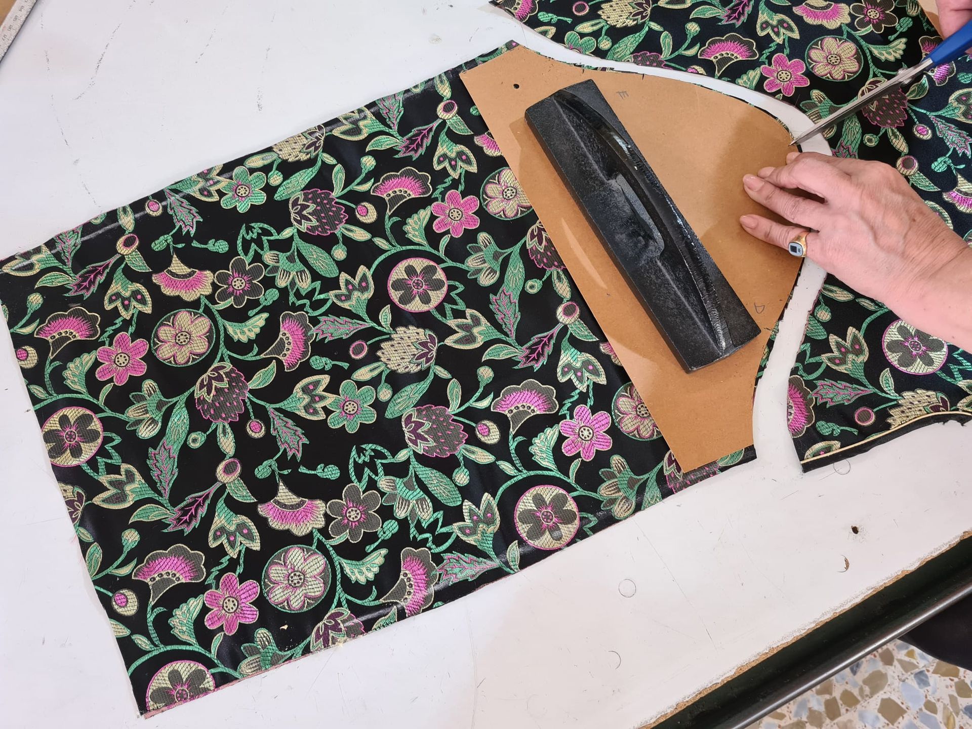 Persona cortando tela floral con tijeras, utilizando una plantilla de papel y un peso negro sobre una superficie blanca.