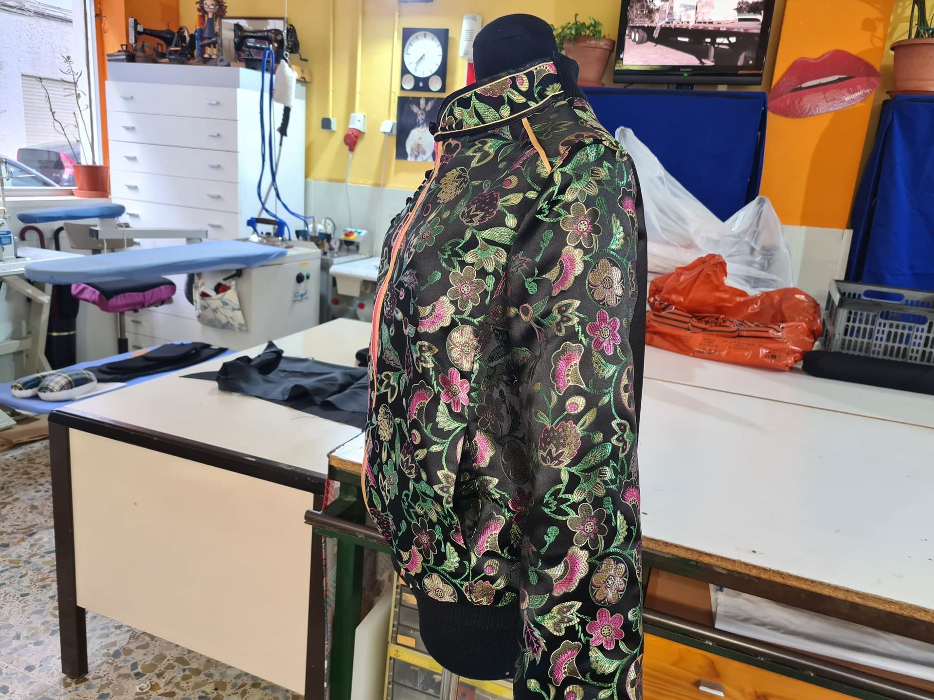 Una chaqueta con estampado floral sobre un maniquí en una sastrería, con máquinas de coser y tablas de planchar visibles.