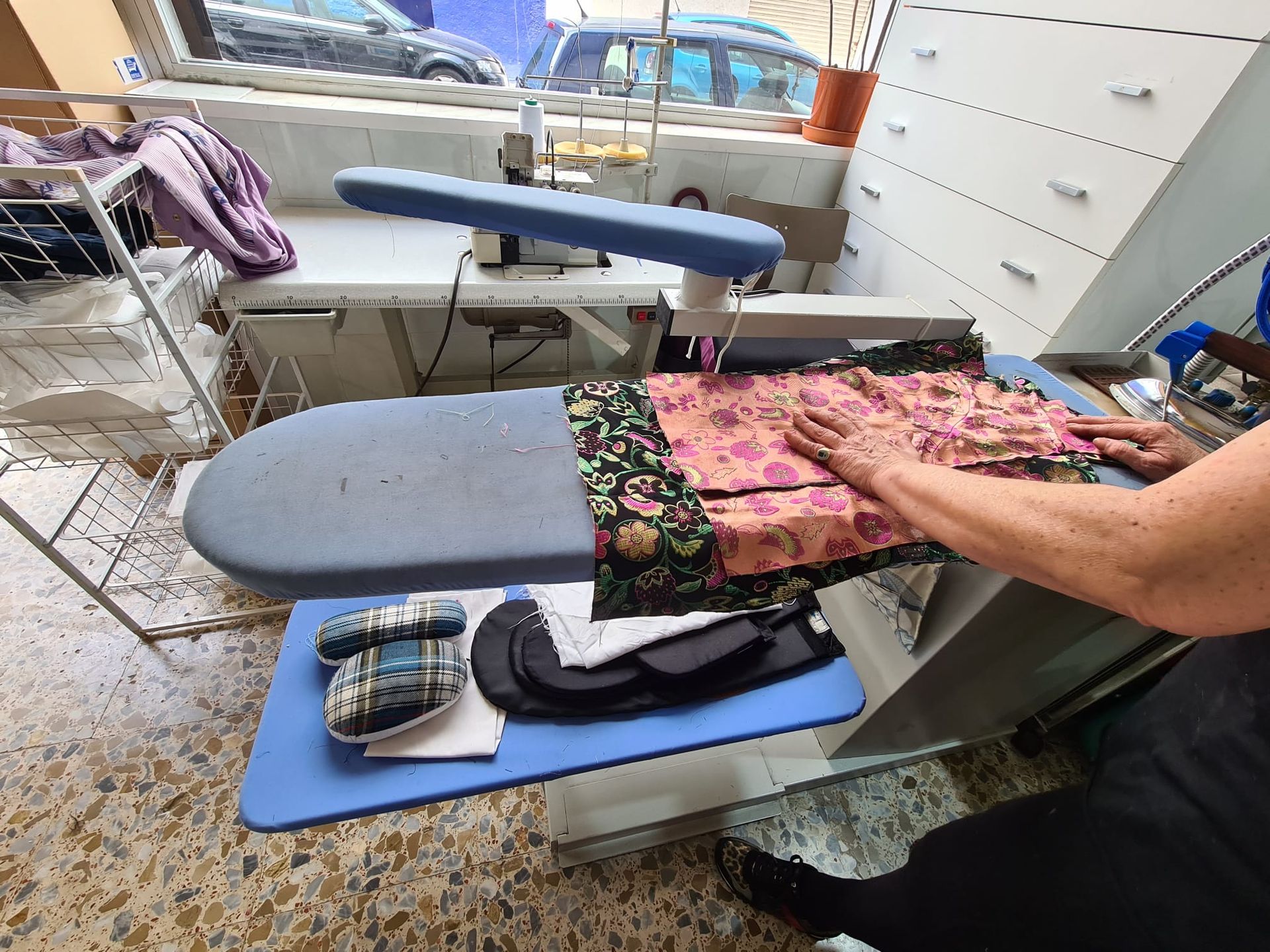 Persona planchando tela en una tabla de planchar. Cuarto de costura con ventana, cajones y tendedero visibles.