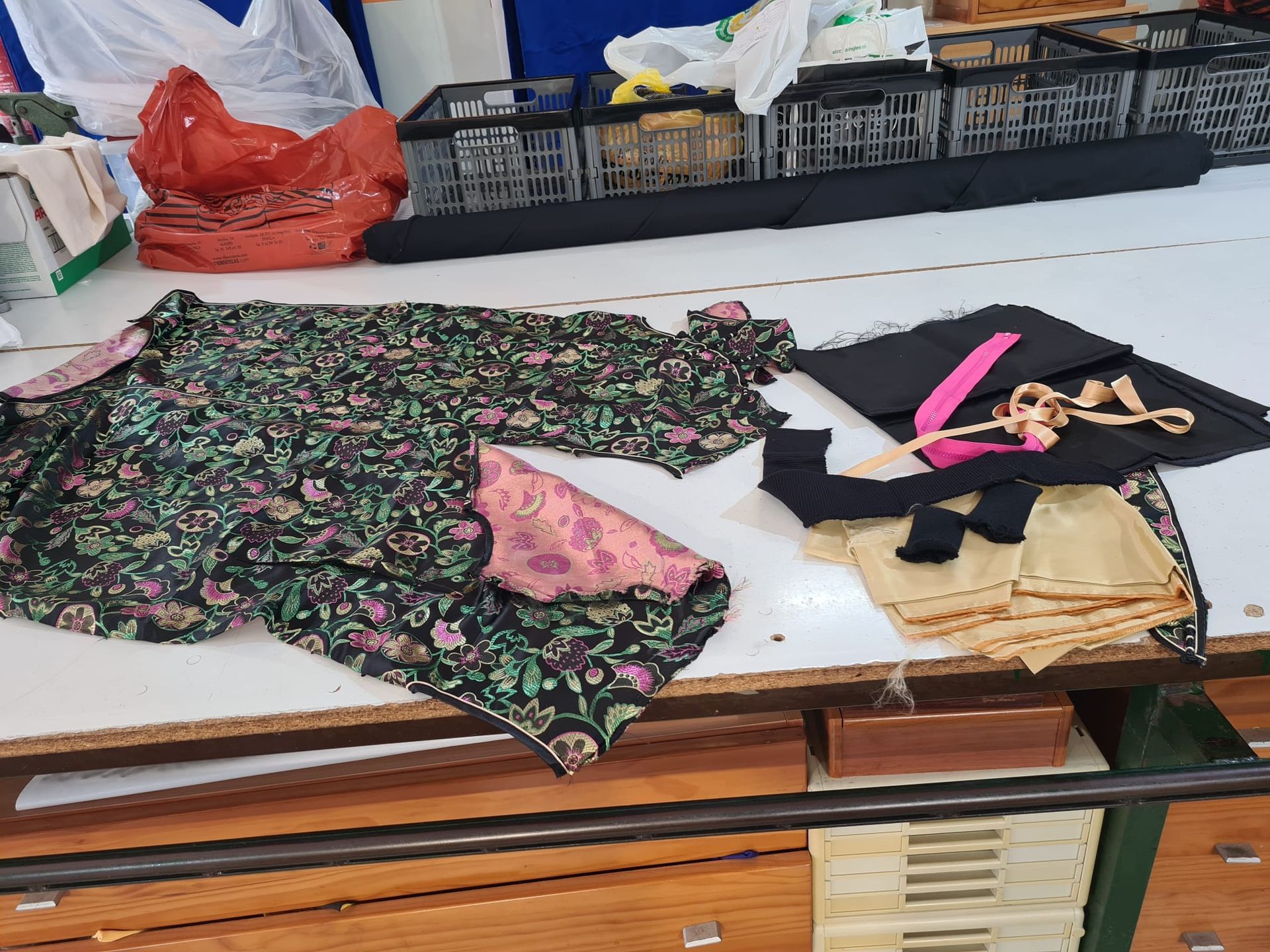 Tela y materiales de costura sobre una mesa blanca. Incluye estampado floral, detalles en rosa, tela negra y ribete dorado.