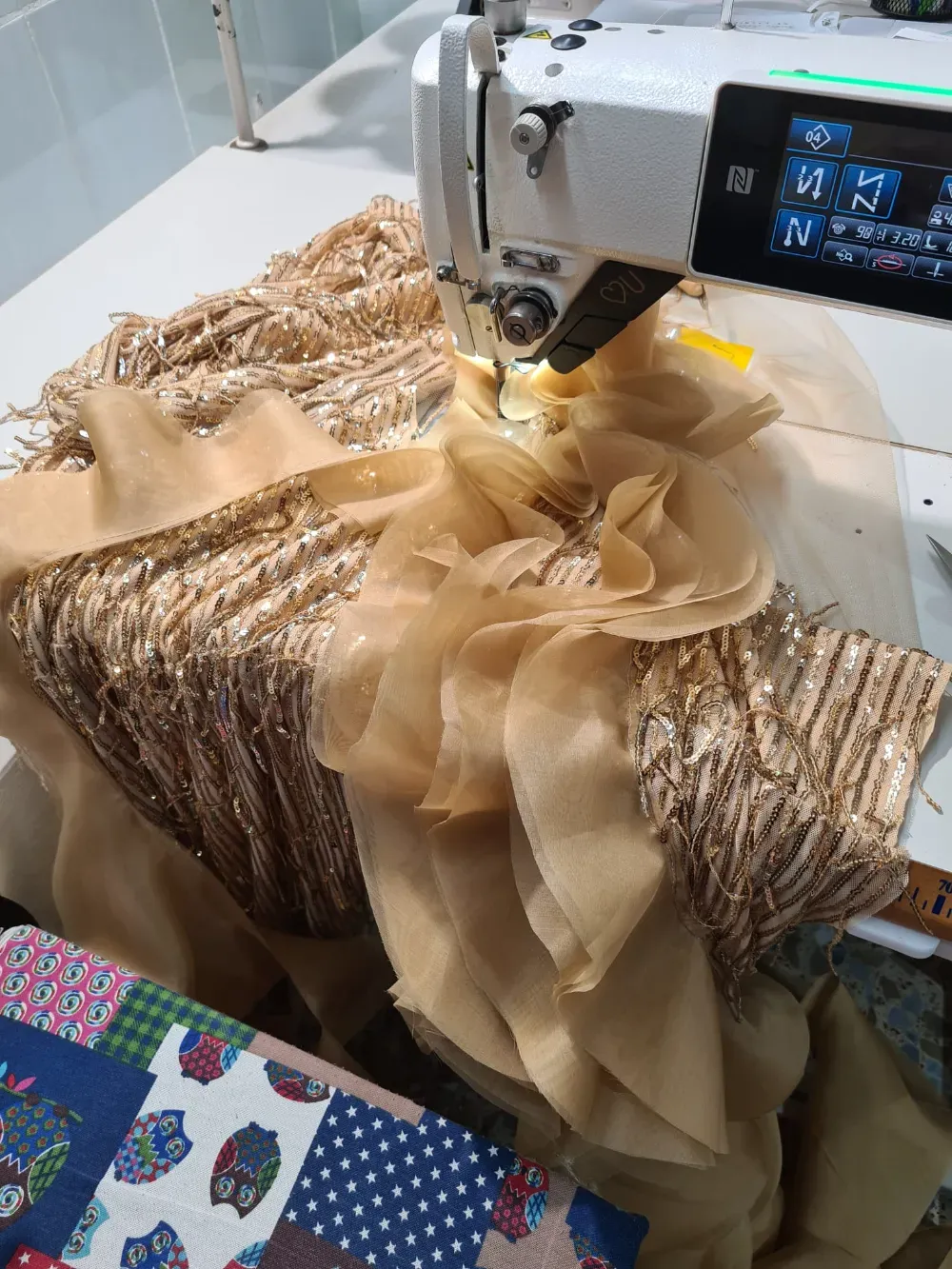 Máquina de coser cosiendo tela con lentejuelas doradas y volantes de gasa sobre una mesa de trabajo.