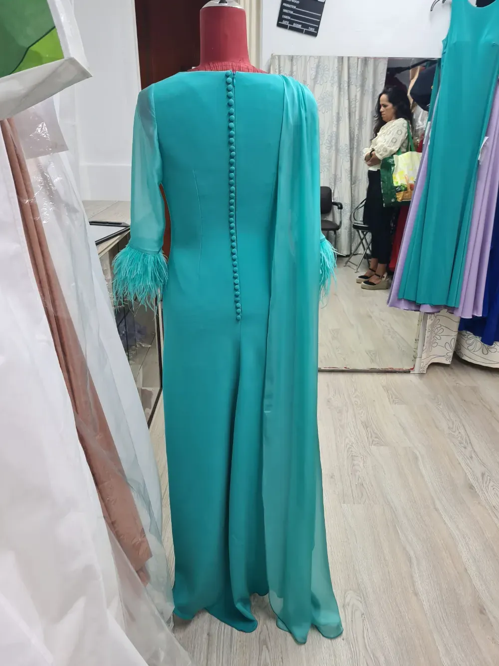Vestido de noche color verde azulado sobre maniquí, mangas largas con puños con plumas, detalle de botones en la espalda y superposición de gasa.