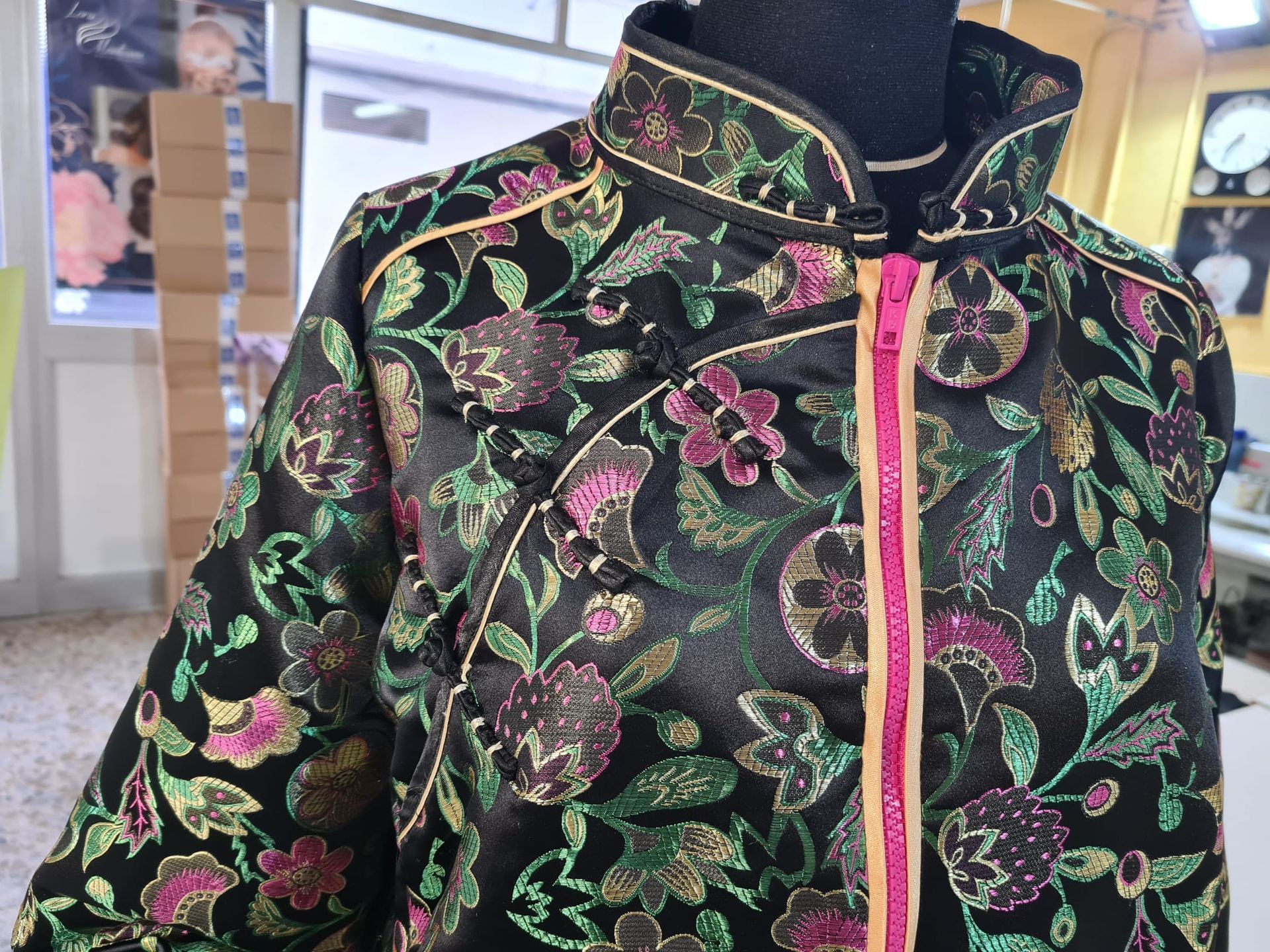 Chaqueta negra con bordado floral, cremallera rosa y ribete dorado.