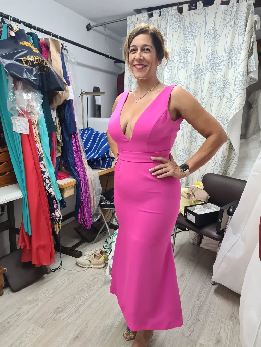 Una mujer con un vestido rosa sonríe, posando en una habitación con percheros y una mesa de costura.
