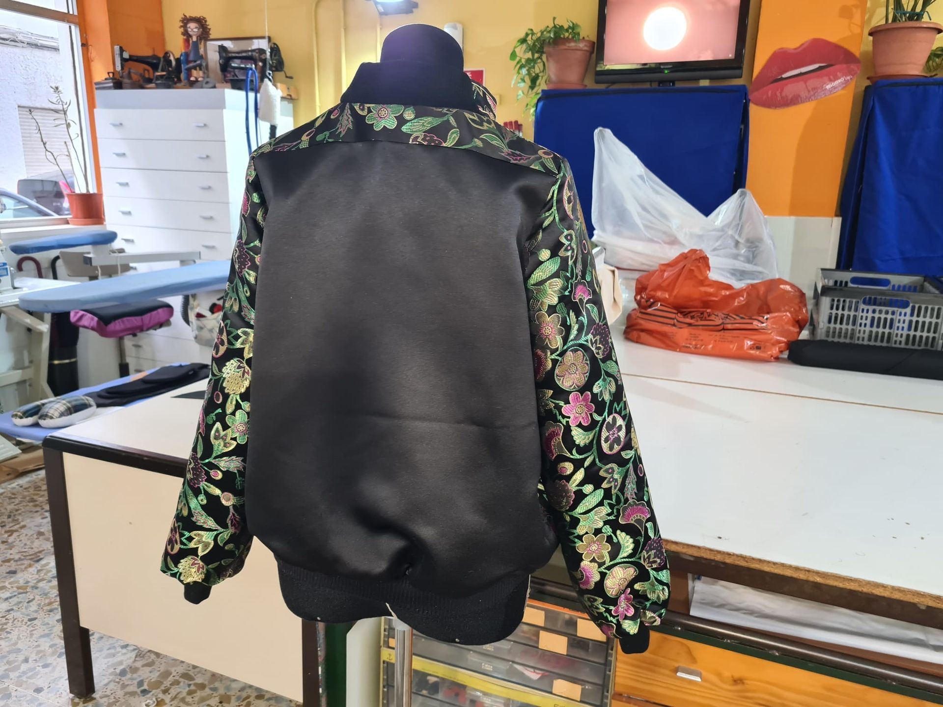 Vista posterior de una chaqueta de satén negra con mangas y cuello con estampado floral, expuesta en un maniquí en un taller.