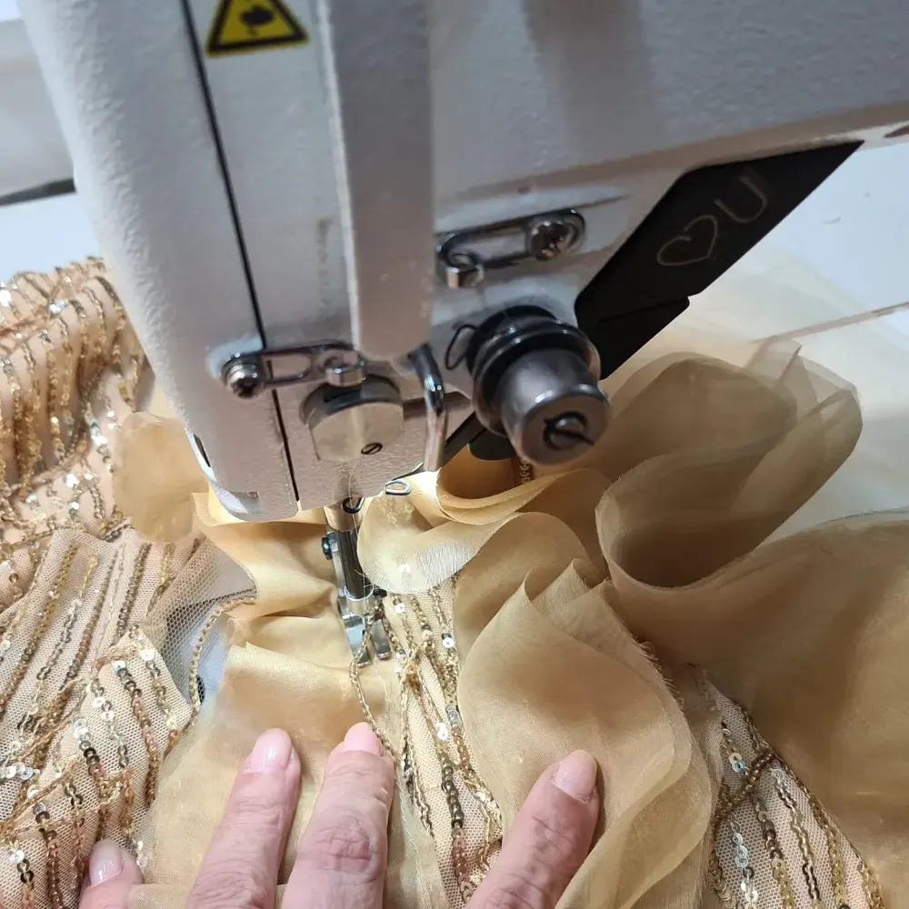 Máquina de coser cosiendo tela dorada con lentejuelas y volantes, guiando el material a mano.